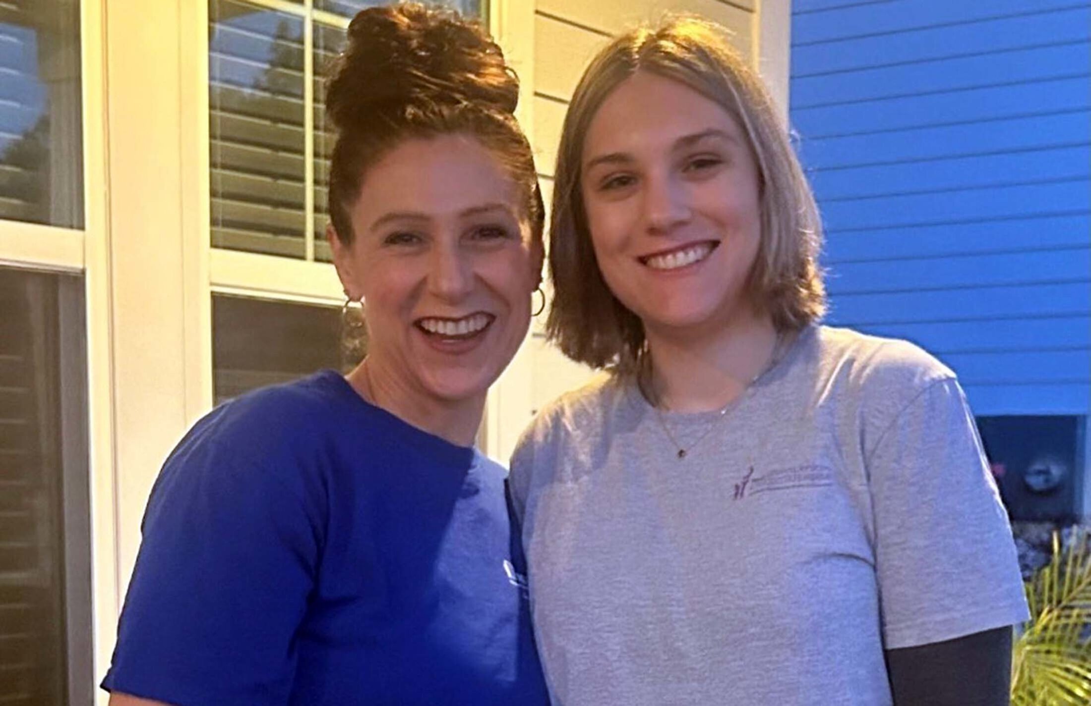 Two women outside, smiling as they lean into each other. The woman on the left has her hair in a bun on top of her head. The woman on the right has shoulder length hair. Both are wearing t-shirts.
