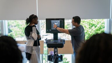 A student holds an ultrasound wand up to the artery in her neck while an instructor points to the screen.