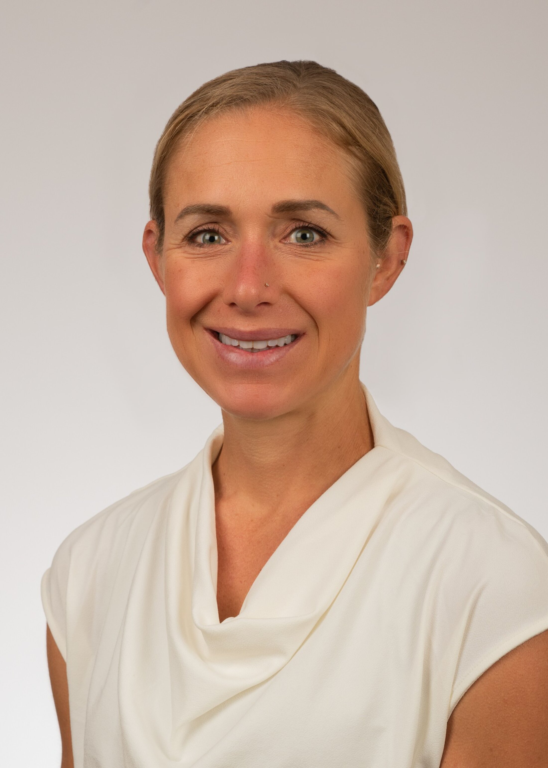 Smiling woman with blonde hair pulled back. She is wearing a sleeveless cream colored top.