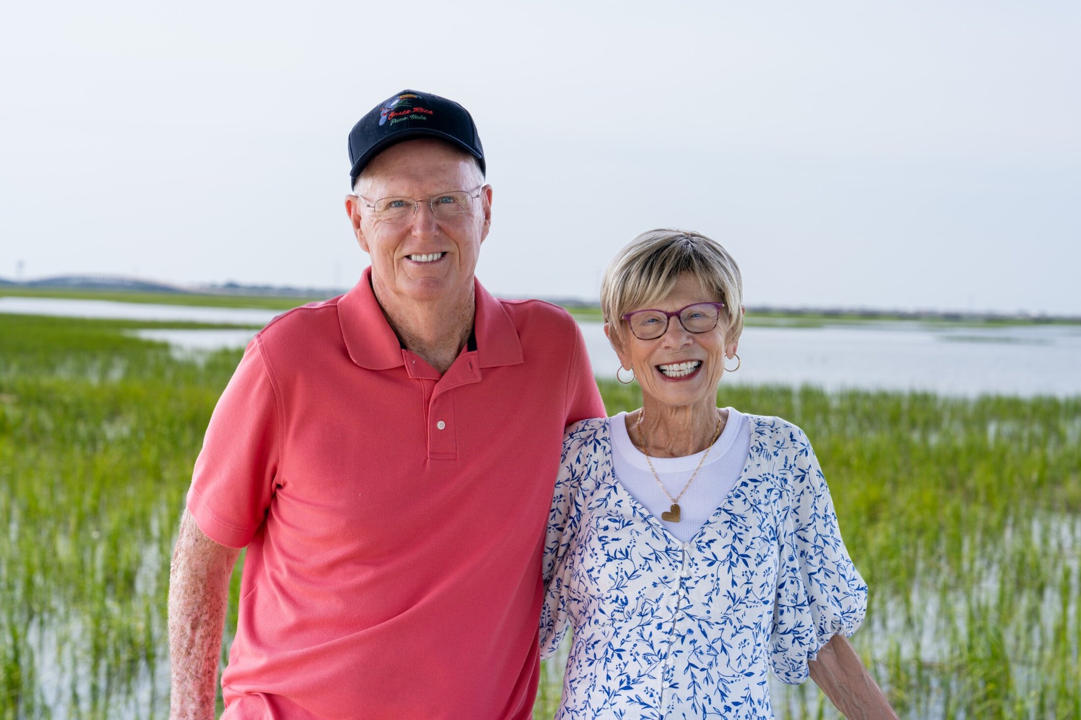 A couple poses in front of a landscape of marsh grass and water extending far into the distance.