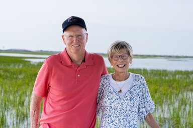 A couple poses in front of a landscape of marsh grass and water extending far into the distance.