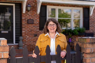 A woman smiles while resting her arms on her picket gate in front of a brick home.