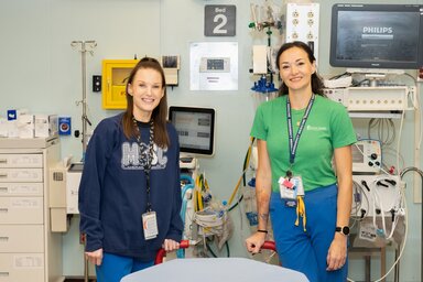 Two women stand side by side in an emergency department compartment. The one on the left has long brown hair pulled back, an MUSC sweatshirt and a lanyard with tags around her neck. The one on the right has black hair pulled back, a green t-shirt that says MUSC health and is also wearing a lanyard with tags. There is a screen that says Philips behind them along with other medical equipment.