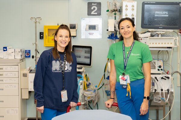 Two women stand side by side in an emergency department compartment. The one on the left has long brown hair pulled back, an MUSC sweatshirt and a lanyard with tags around her neck. The one on the right has black hair pulled back, a green t-shirt that says MUSC health and is also wearing a lanyard with tags. There is a screen that says Philips behind them along with other medical equipment.