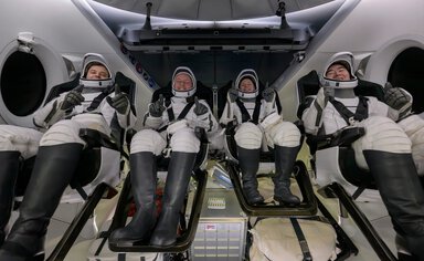 Four people in space suits give thumbs up to camera.