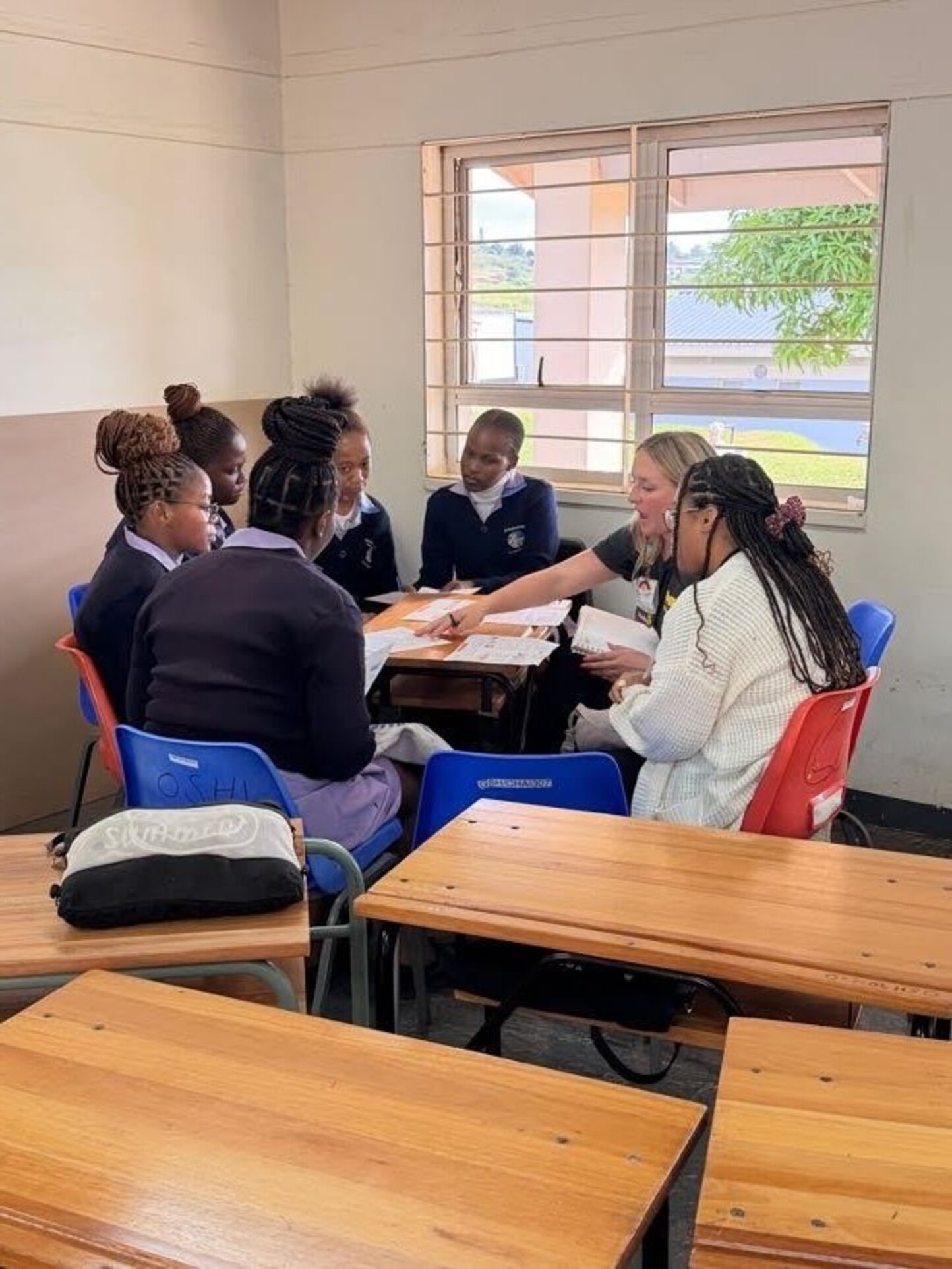 A woman sits at a desk with a group of five students in school uniforms, reviewing papers together in a classroom setting.
