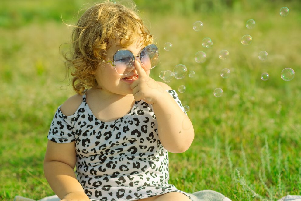 A cheerful child dressed in a leopard print shirt is playing outside The sun shines brightly as the kid adjusts their sunglasses and smiles surrounded by green grass and trees