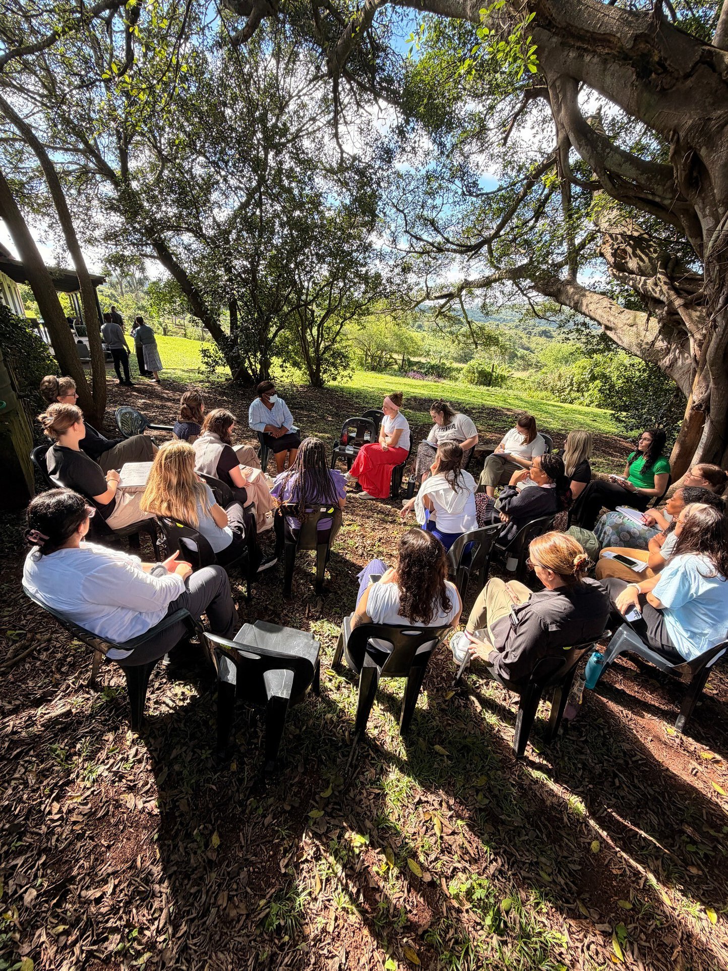 A large group of people sit in a circle of plastic chairs outdoors under large trees, engaged in a discussion or seminar, with rolling green hills visible in the background.