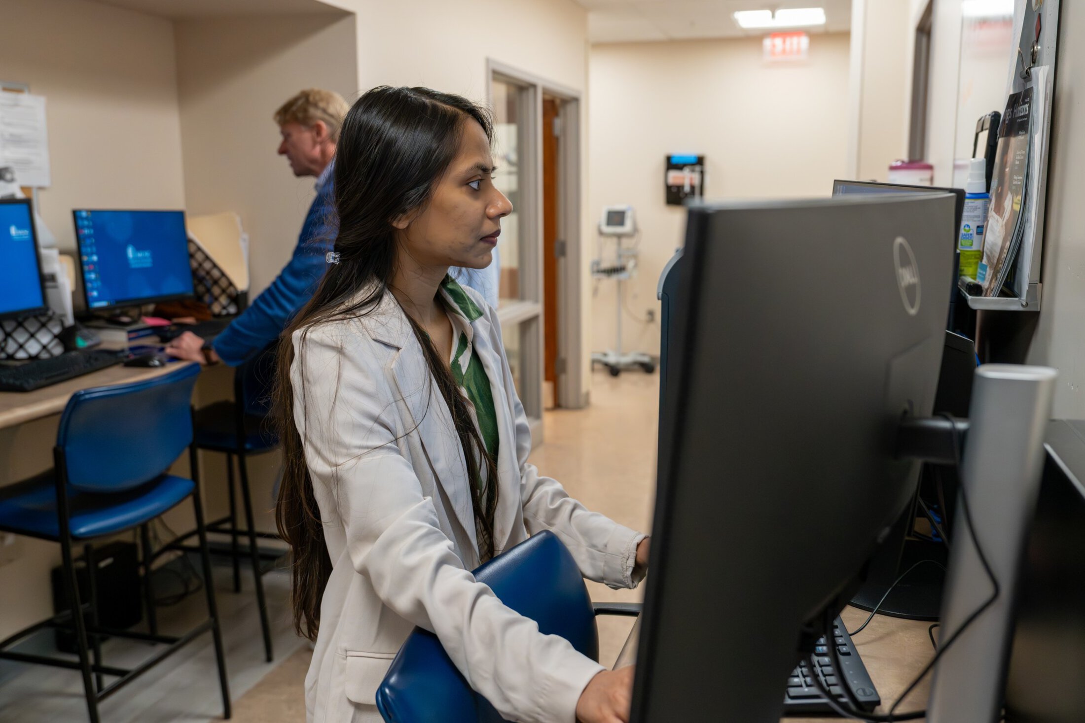 Two doctors each look at different computers in a work area outside clinic exam rooms.