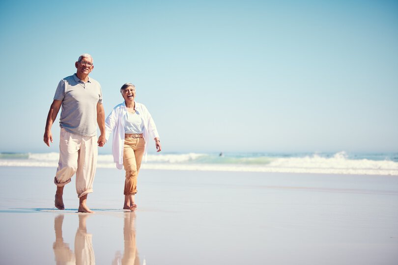 Holding hands, summer and an old couple walking on the beach with a blue sky