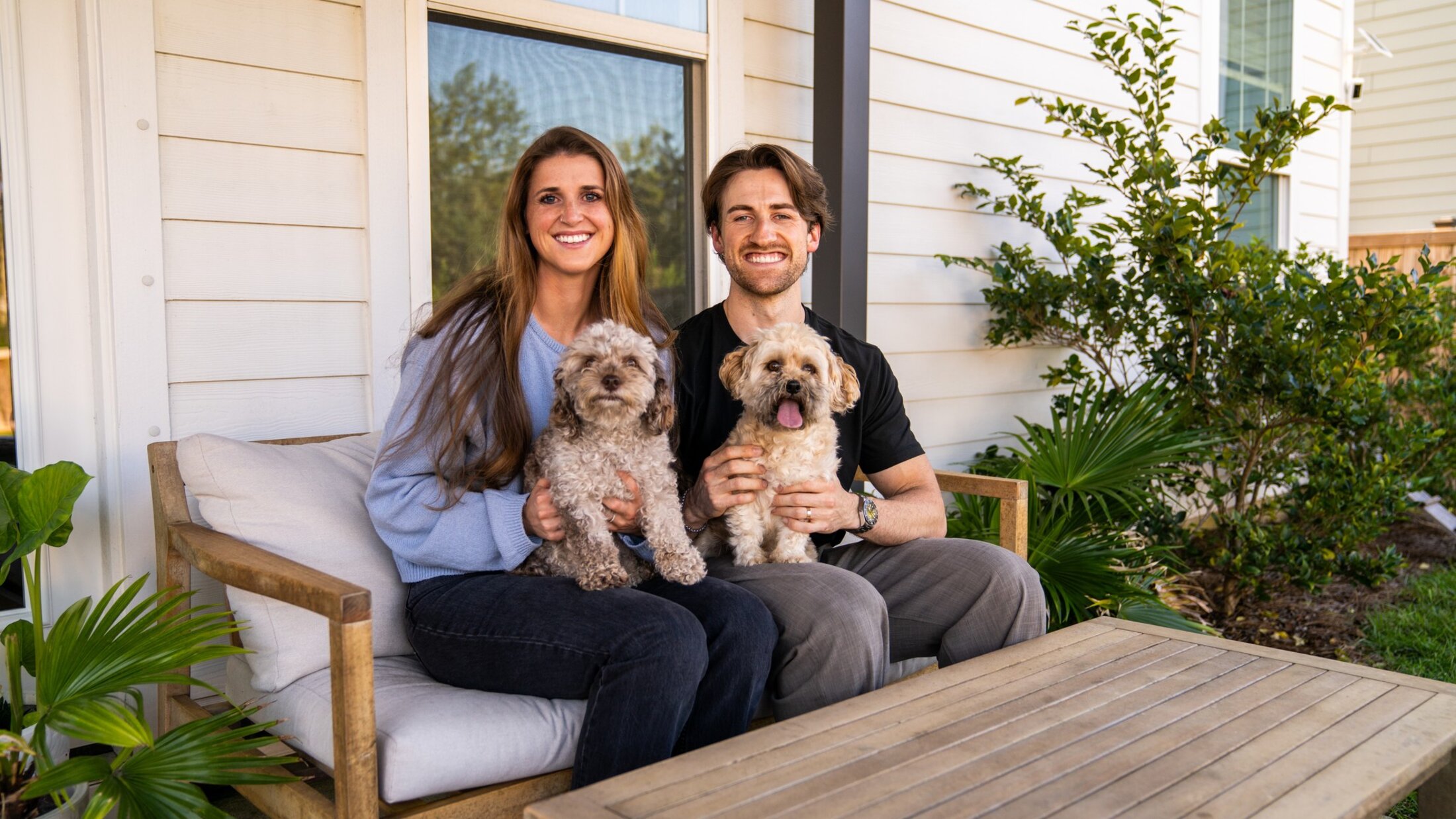 A man and woman sit on a bench in their backyard with two small fuzzy white dogs on their laps.