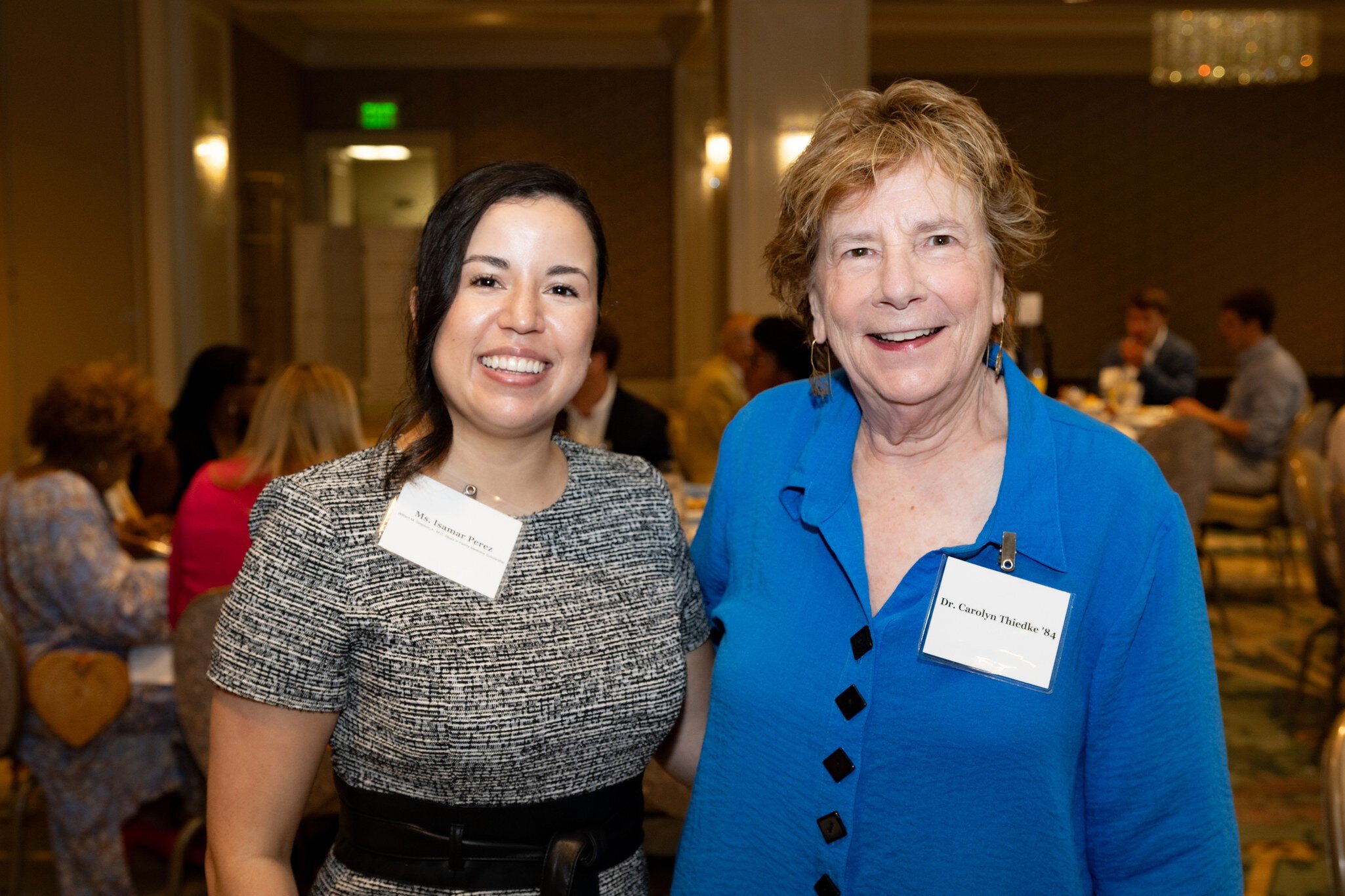 Carolyn Thiedke and Isamar Perez Rodriguez smile at the camera at a formal event. One wears a gray dress, the other a blue blouse. People are seated in the background, creating a warm, friendly atmosphere.