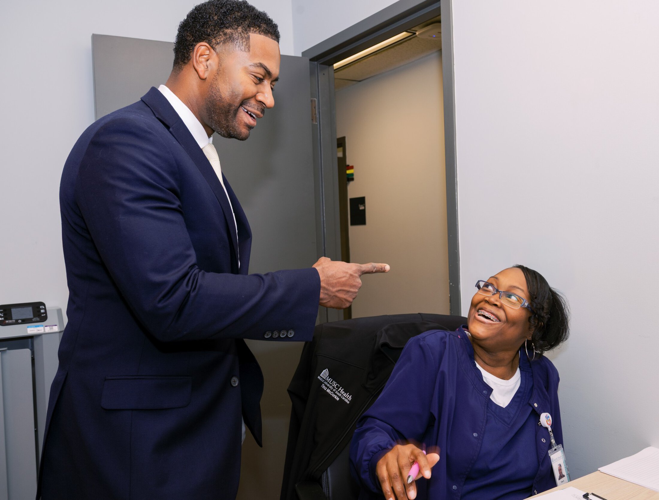 A woman wearing hospital scrubs and glasses smiles as she looks up at a man who is gesturing to her. He is wearing a suit and smiling as well.