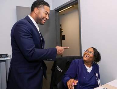 A woman wearing hospital scrubs and glasses smiles as she looks up at a man who is gesturing to her. He is wearing a suit and smiling as well.