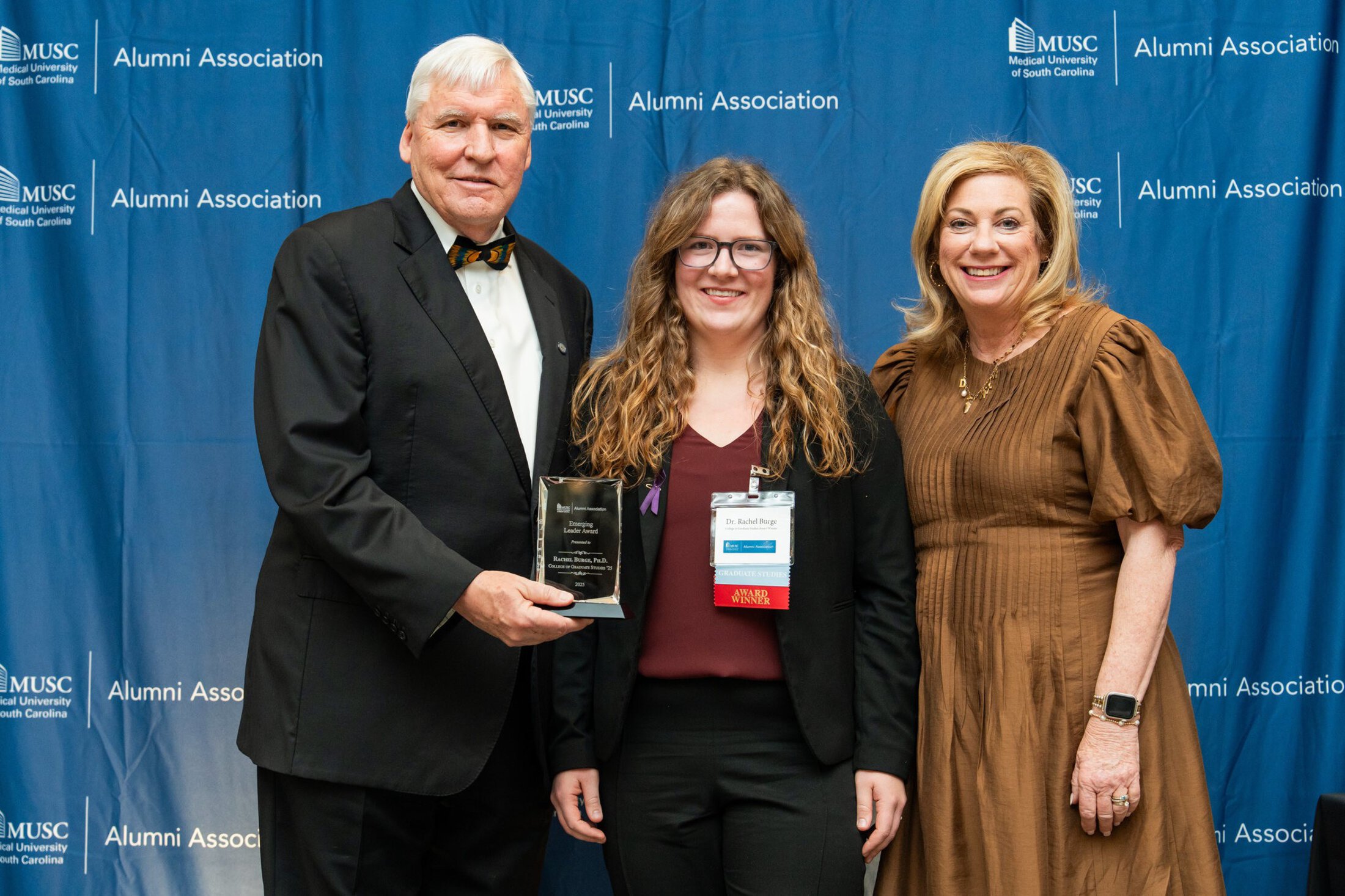 Rachel Burge stands with MUSC president David Cole and first lady Kathy Cole while receiving the Emerging Leader Award at an MUSC Alumni Association event. She holds a glass award and smiles in front of a blue MUSC Alumni Association backdrop.