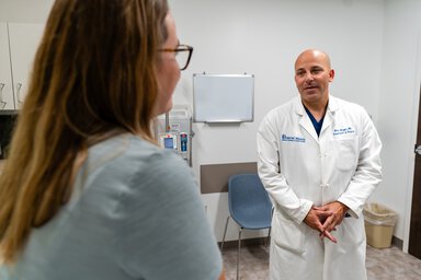 A doctor in a white coat speaks with a nurse in a clinic exam room.