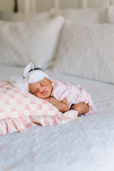 Tiny baby with a white bow in her hair wearing a pink outfit sleeps on a pink and white checked pillow.