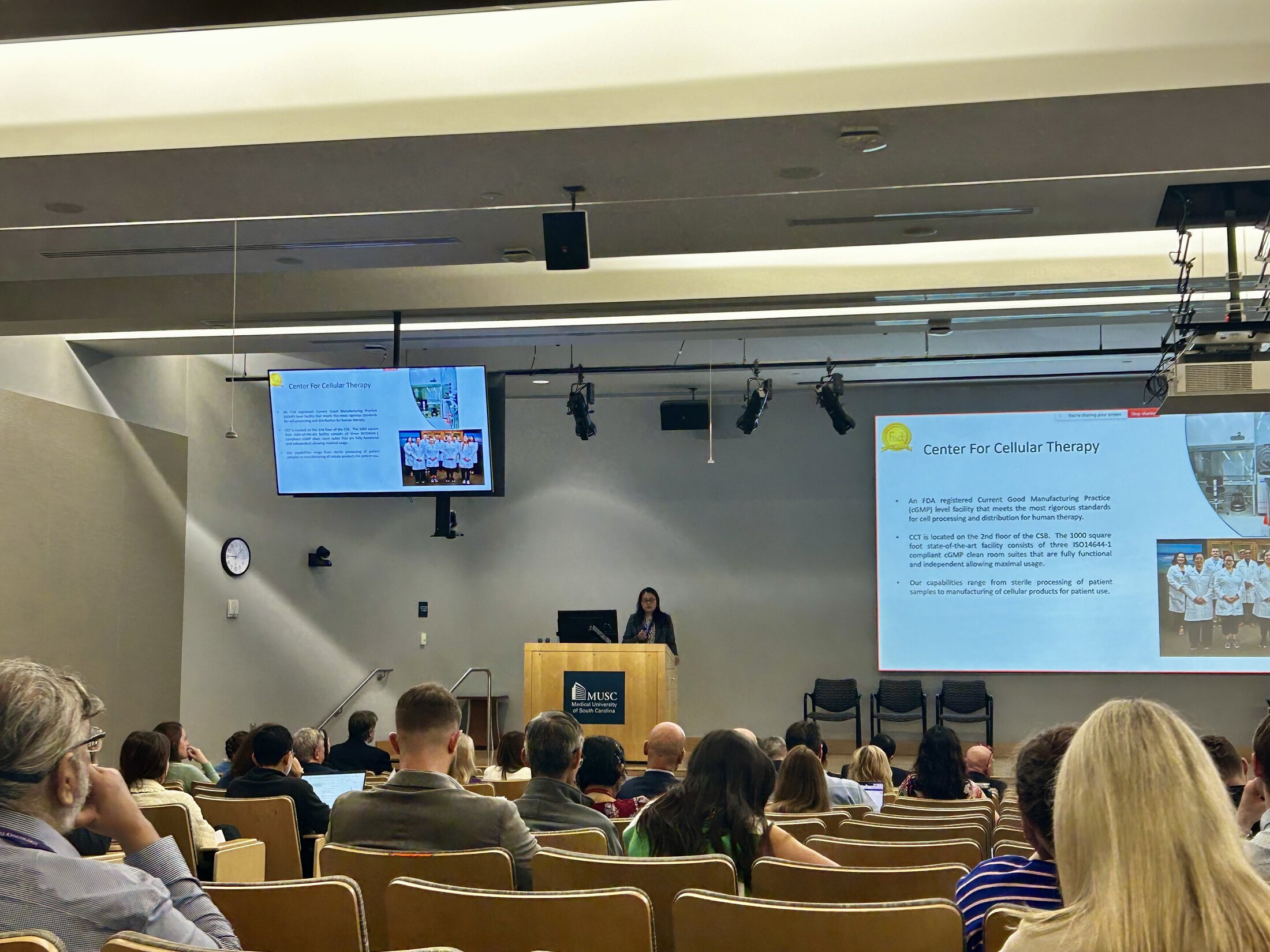 A speaker presents at a podium in a large auditorium at MUSC, addressing an audience seated in tiered rows. Two screens display a presentation slide titled “Center for Cellular Therapy,” with images of a lab and staff.