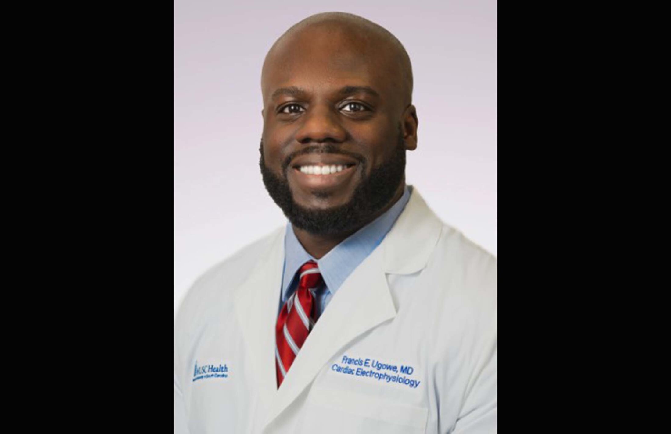 Dr. Francis Ugowe smiles while wearing a white doctor's coat and a necktie.