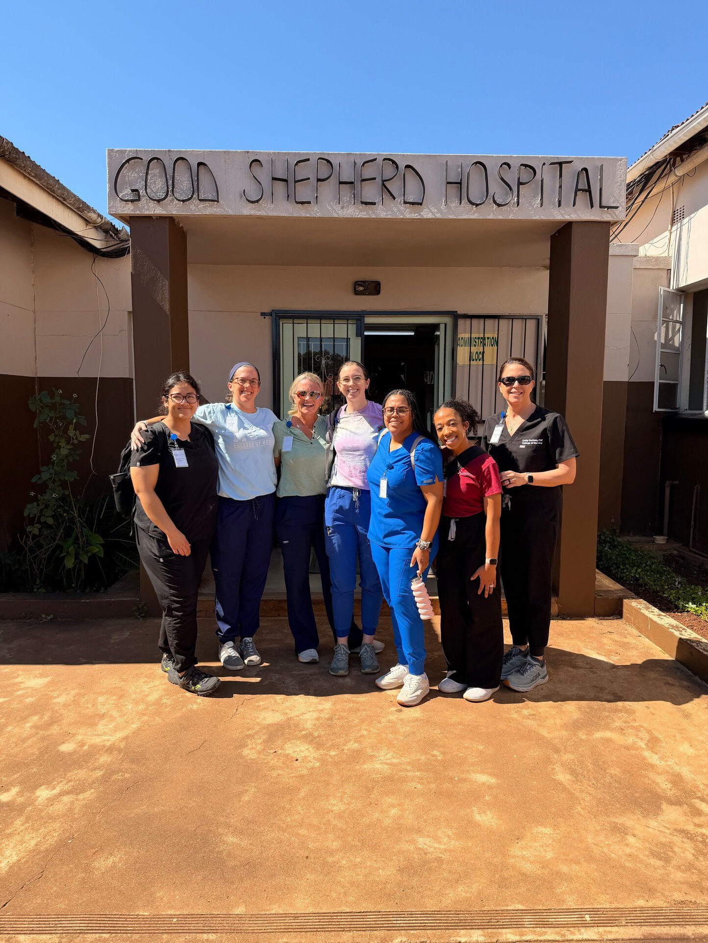 Seven women in scrubs and casual clothes stand smiling in front of a building with a concrete sign reading Good Shepherd Hospital.