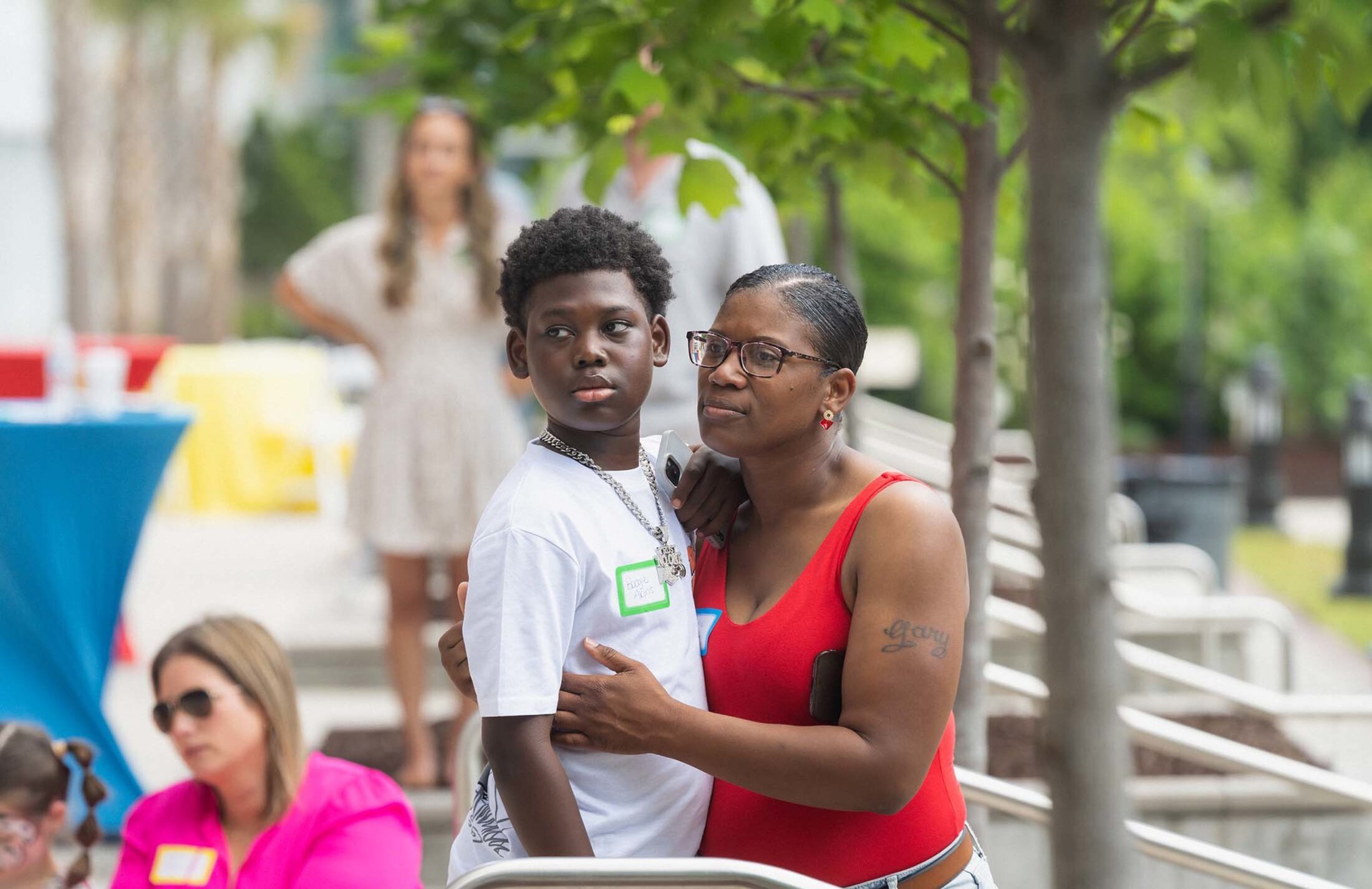 A young man stands with a woman has her arms around him. They are looking to the side, appearing to listen to someone speak. He is wearing a white t-shirt. She is wearing a red top.