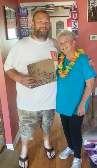 A man holds a sign that says welcome home. He's wearing a white t-shirt and camo shorts. His arm is around a woman who is wearing a lei, a blue t-shirt an black pants. There are patriotic signs behind them in a hallway of a house.