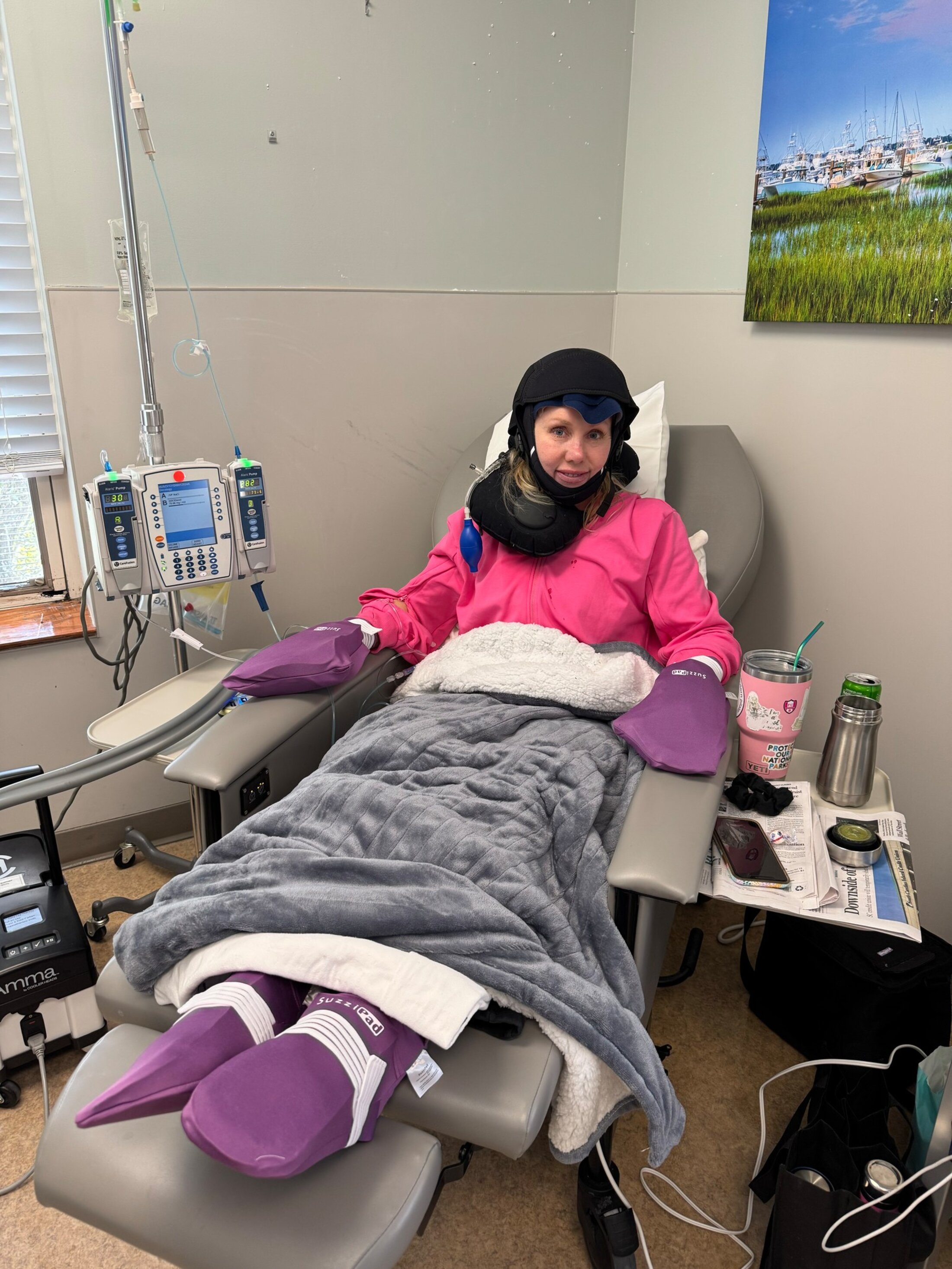 A woman sits in an infusion chair with oversized mitten-like contraptions on her hands and feet and a shawl-like cap wrapped around her head and neck.