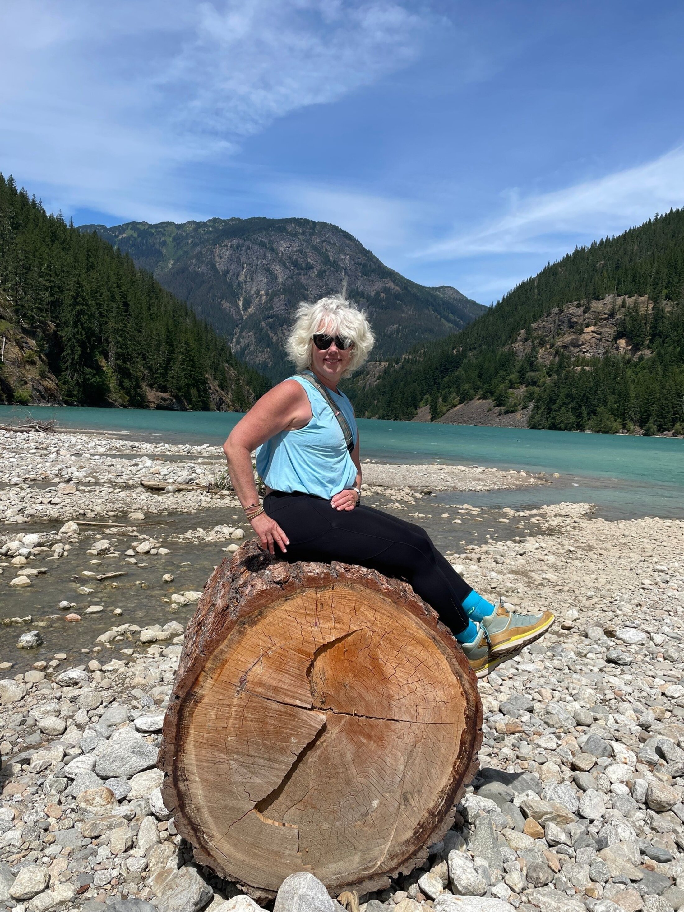 A woman sits perched on a fallen log on a rocky riverbed with a sea-glass green lake and pine-covered mountains behind her.