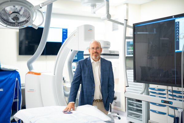 A smiling man wearing a blue jacket and white shirt stands in front of large white devices. There is a screen on the right side of the image. It appears to be an operating room.
