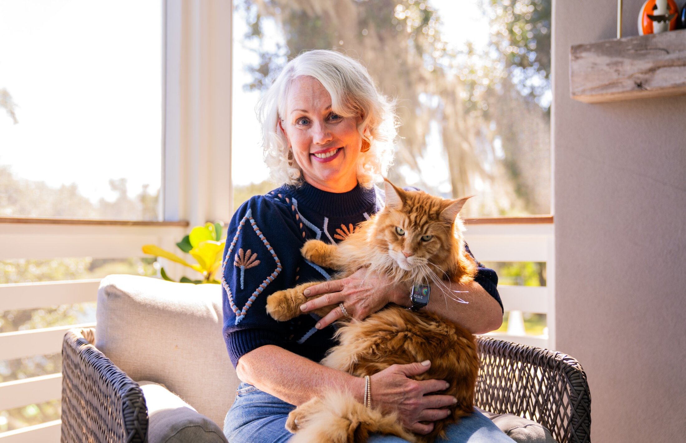 A woman sits near a sunny window holding an enormous Maine coon cat, who glowers at the camera.
