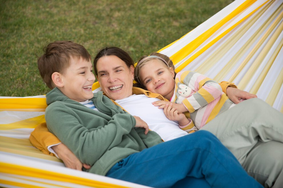 A mom lays back in a hammock, snuggling a boy and girl on either side of her, laughing.