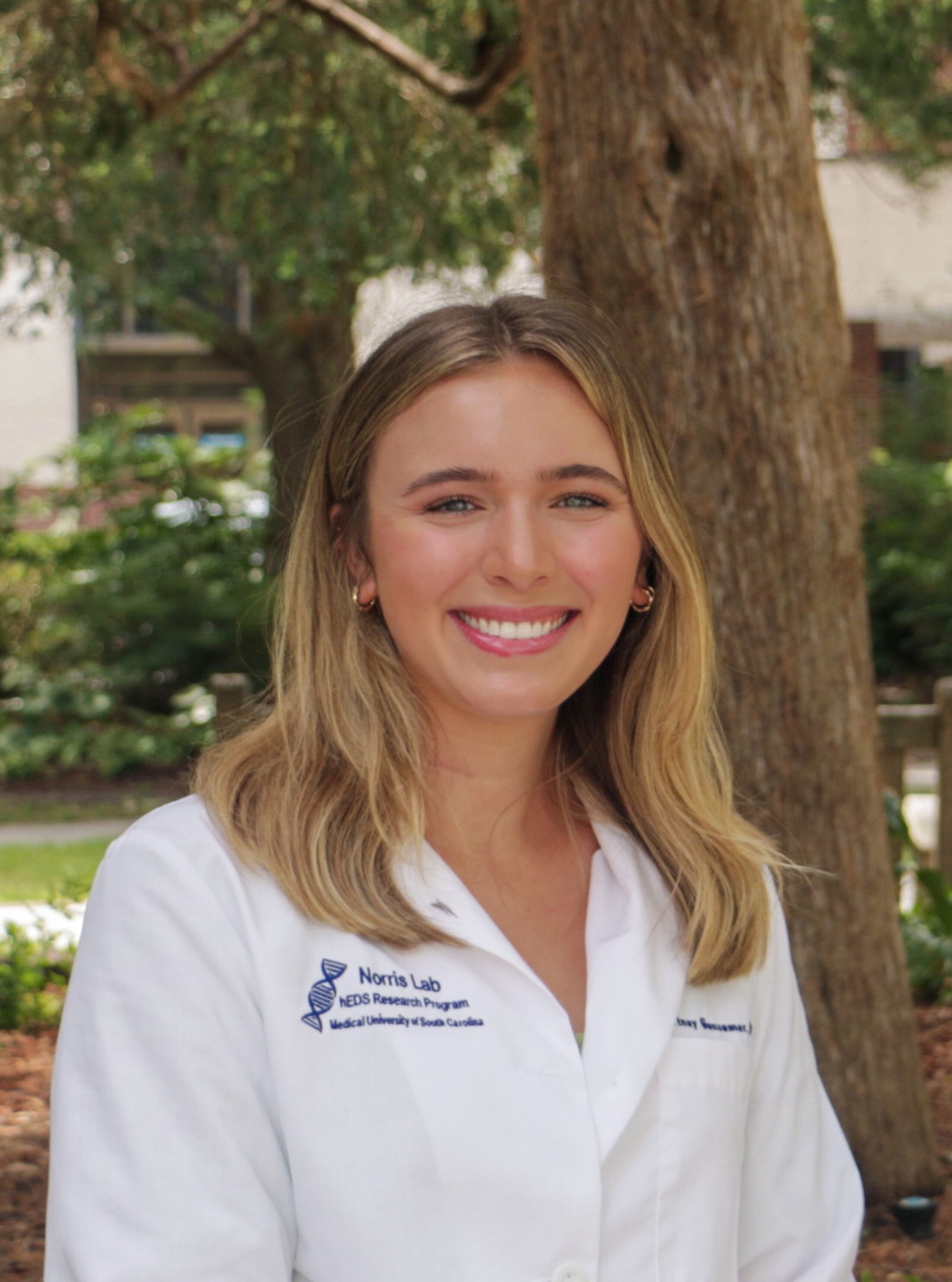 Young woman with shoulder-length blonde hair smiling at the camera while wearing a white research lab coat outdoors, with trees and greenery in the background.