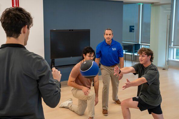 A man instructs three male students through an exercise with a fitness ball.