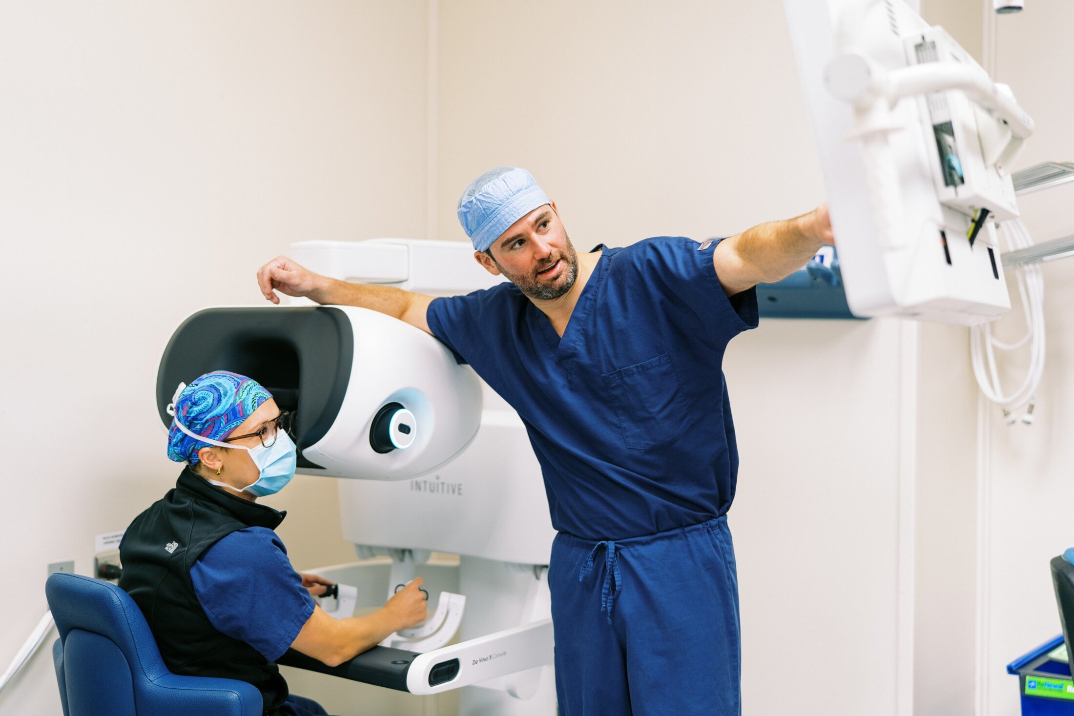 Ben Stone, M.D., gestures to a screen that magnifies the robotic surgery that urology resident Jennifer Drerup, M.D., seated at the robot controls, is performing.