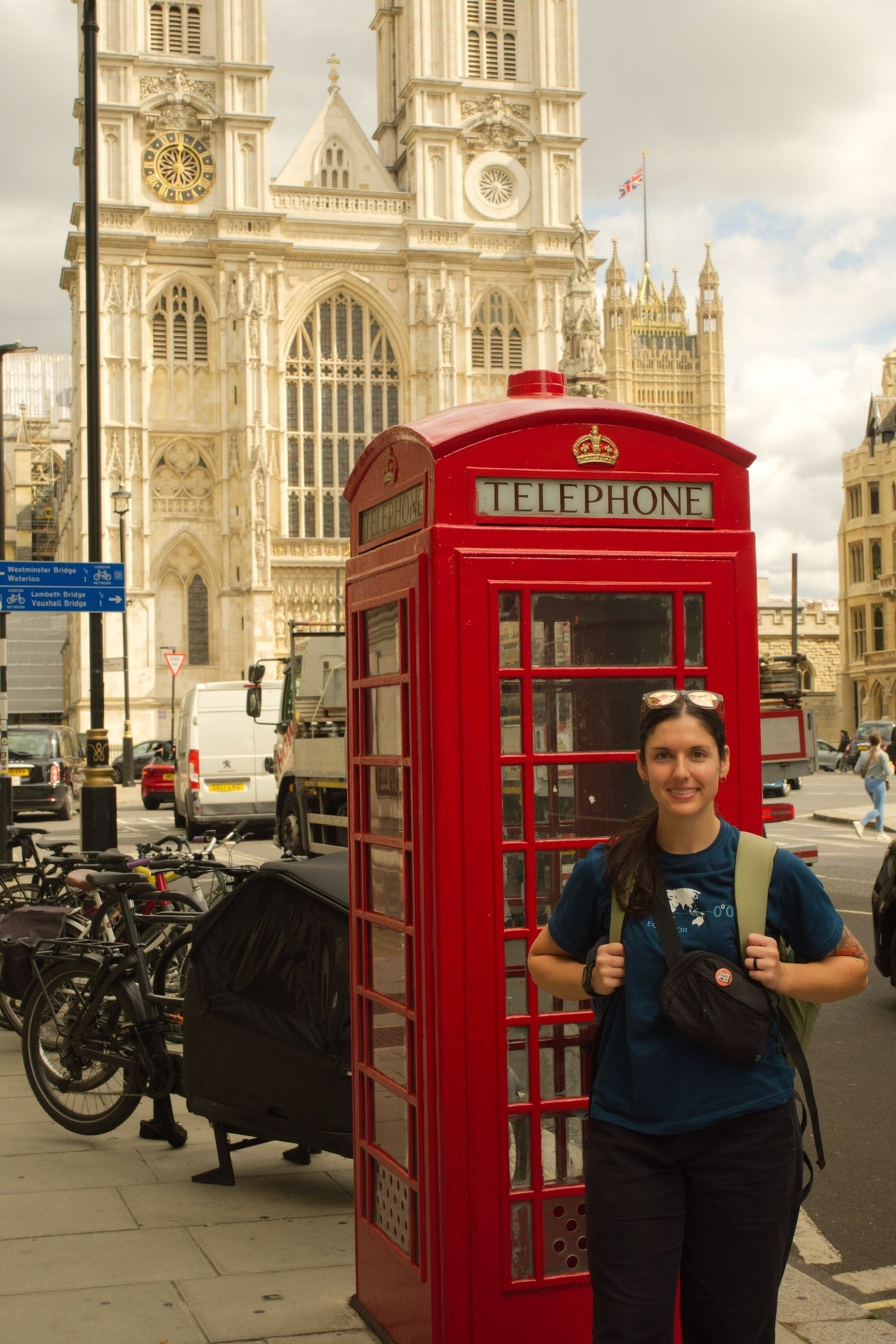 Young woman wearing navy t-shirt and a backpack stands in front of a red telephone booth. There is an impressive building behind her in London.