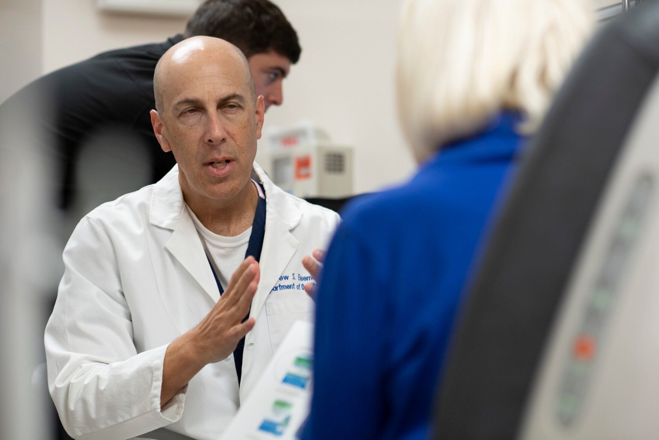 Man wearing a white lab coat gestures as he speaks to another person whose back is to the camera. A third person is behind the speaker leaning over to look at something.