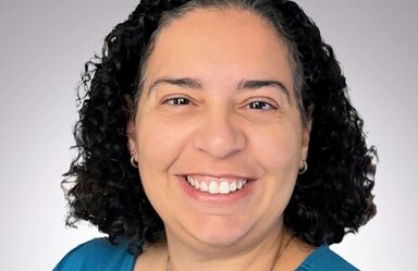 Headshot of smiling woman with curly dark hair. She's wearing earrings, a necklace and a blue top.