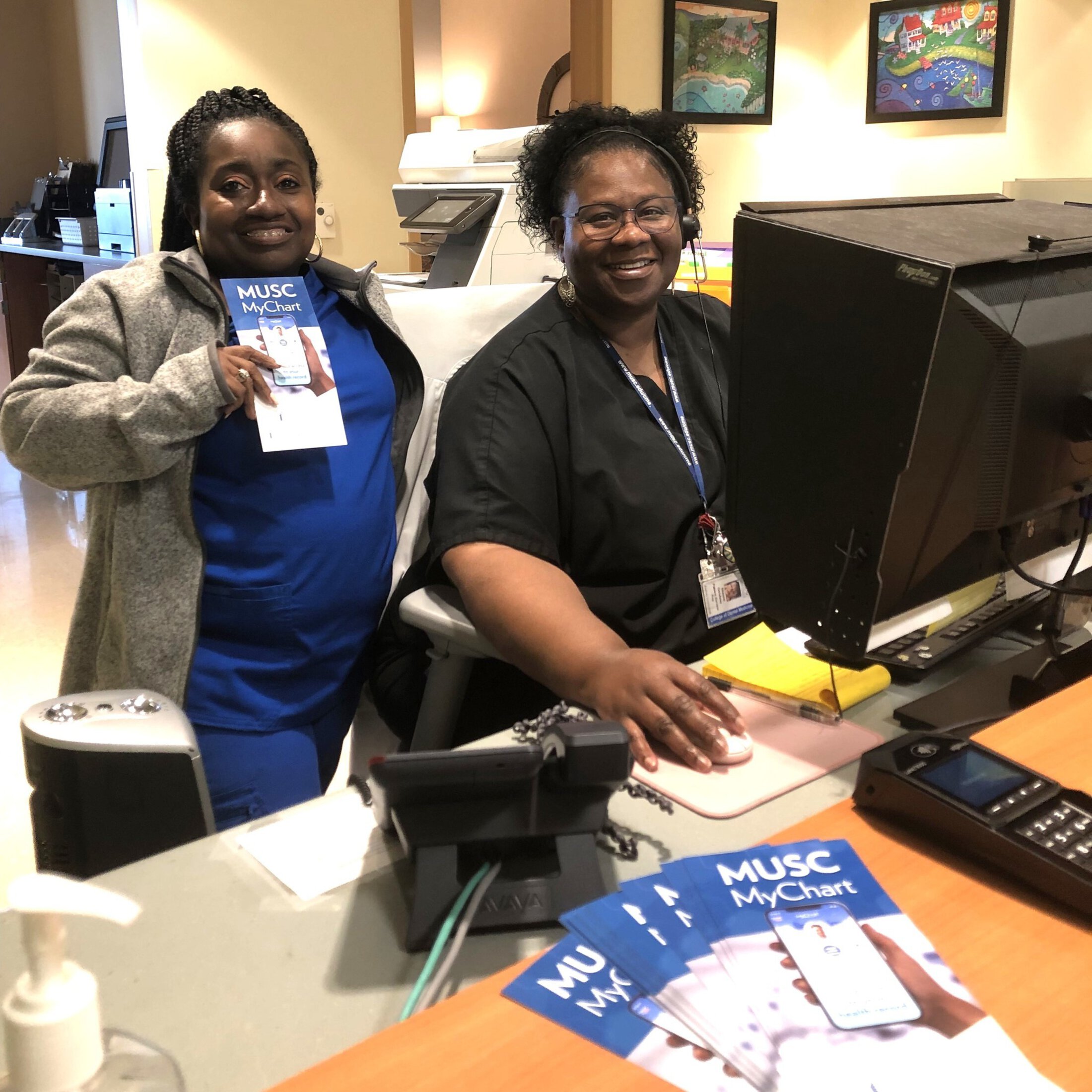 Two front desk employees at the MUSC dental clinic smile at the camera. One is holding a “MUSC MyChart” pamphlet.