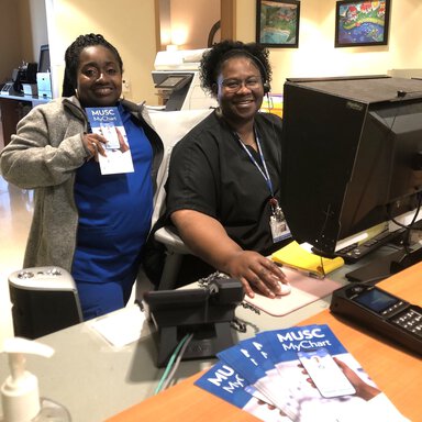 Two front desk employees at the MUSC dental clinic smile at the camera. One is holding a “MUSC MyChart” pamphlet.