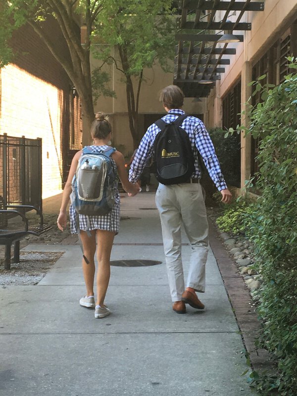 A couple in matching checkered outfits and backpacks walk hand in hand down a tree-lined path, conveying a sense of togetherness and harmony.