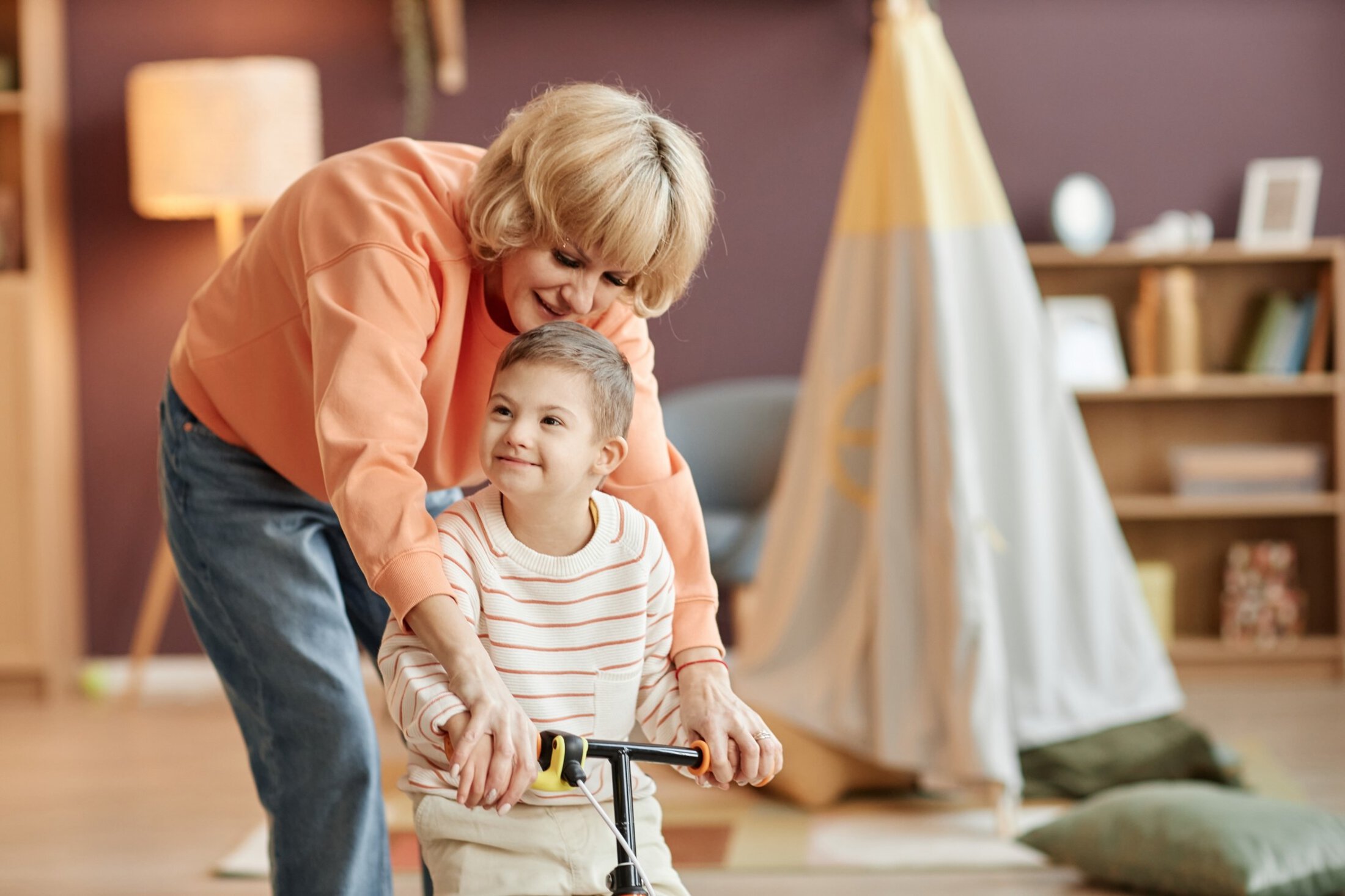 MUSC Health Childrens In Patient Rehab Portrait of happy mother helping boy with down syndrome using runbike indoors