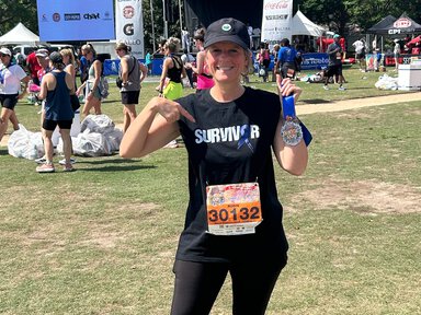 a woman holds up a 10K medal and points to the word 'survivor' on her T-shirt