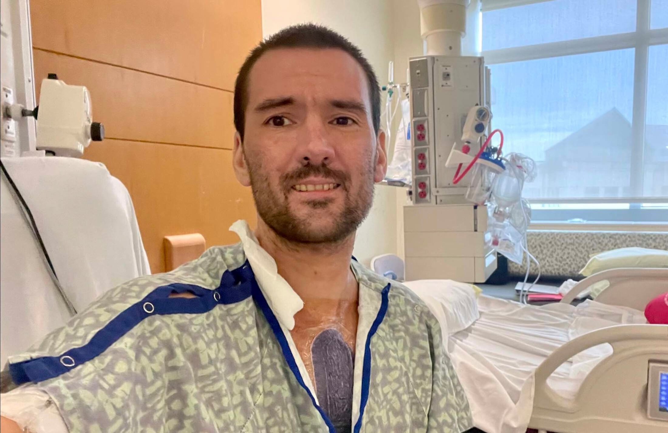 A man with dark hair and a goatee is in a hospital room. He's wearing a gown that is open at the chest, which reveals a large dark colored covering over his chest.