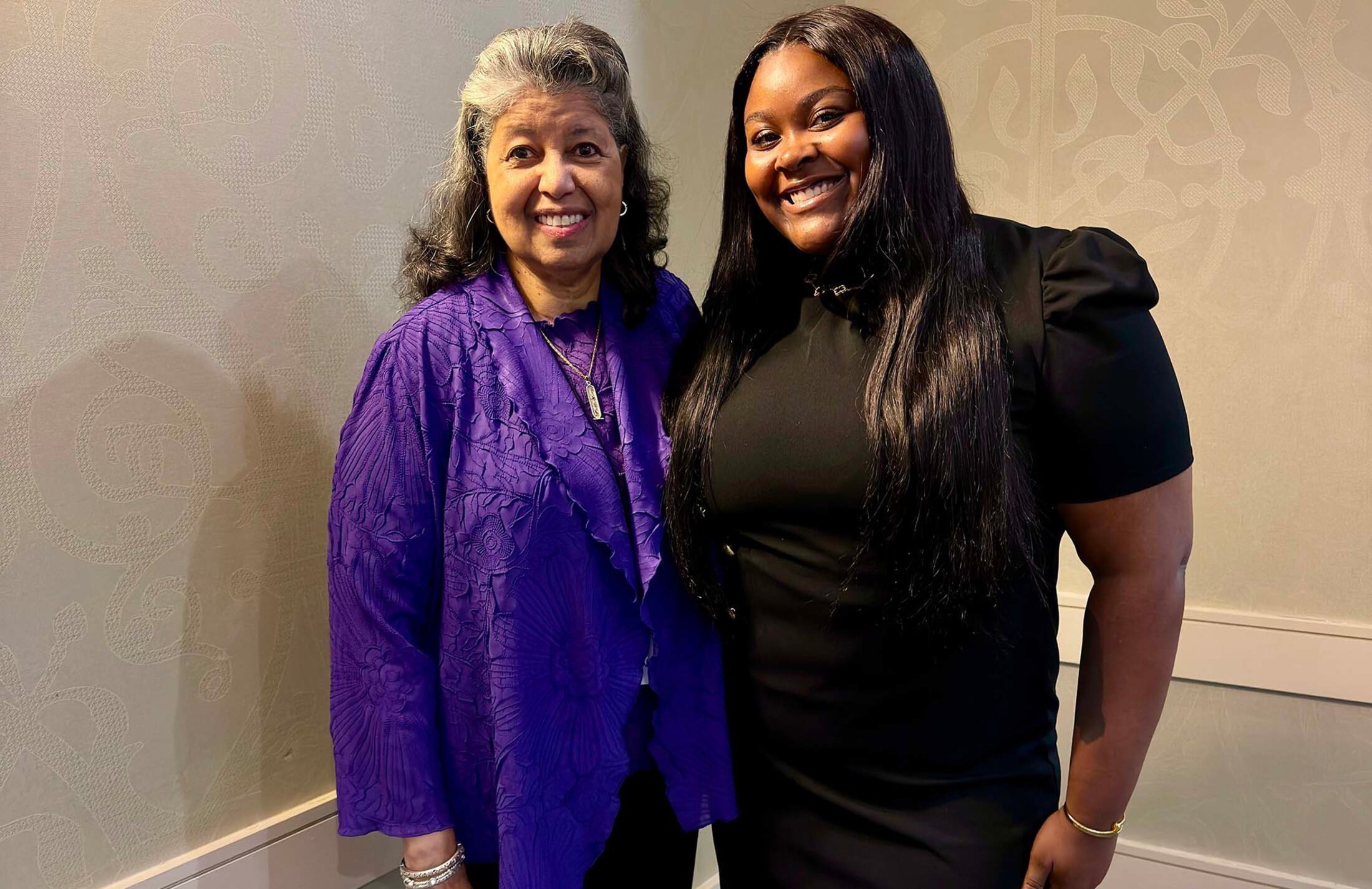 A woman wearing a purple top and jacket smiles. She is standing with a younger woman. The younger woman has long dark hair and is wearing a black dress with short sleeves.