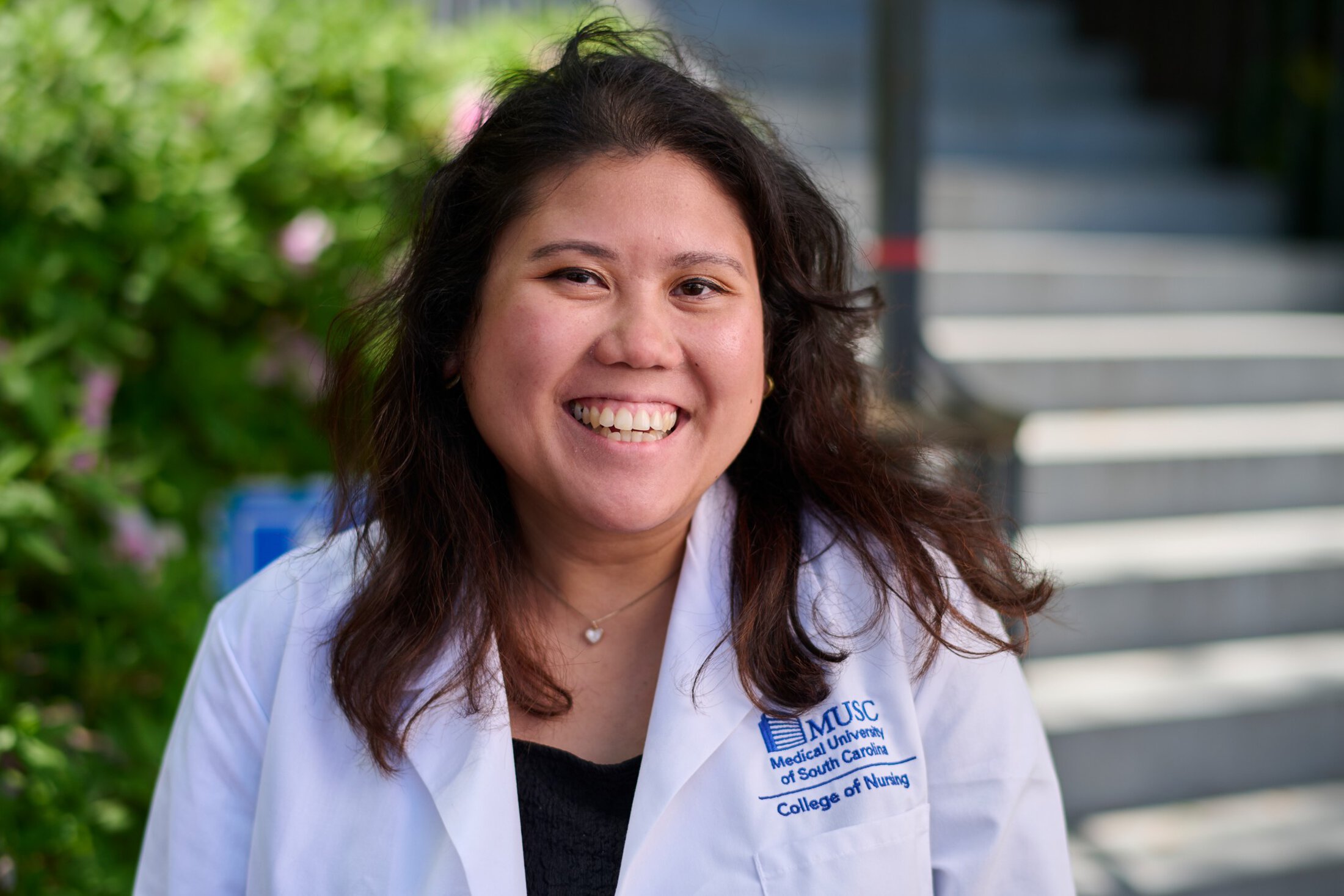 Portrait of Danielle Mercado, MUSC College of Nursing, smiling outdoors while wearing a white MUSC College of Nursing coat, outdoors with greenery and campus steps softly blurred in the background.