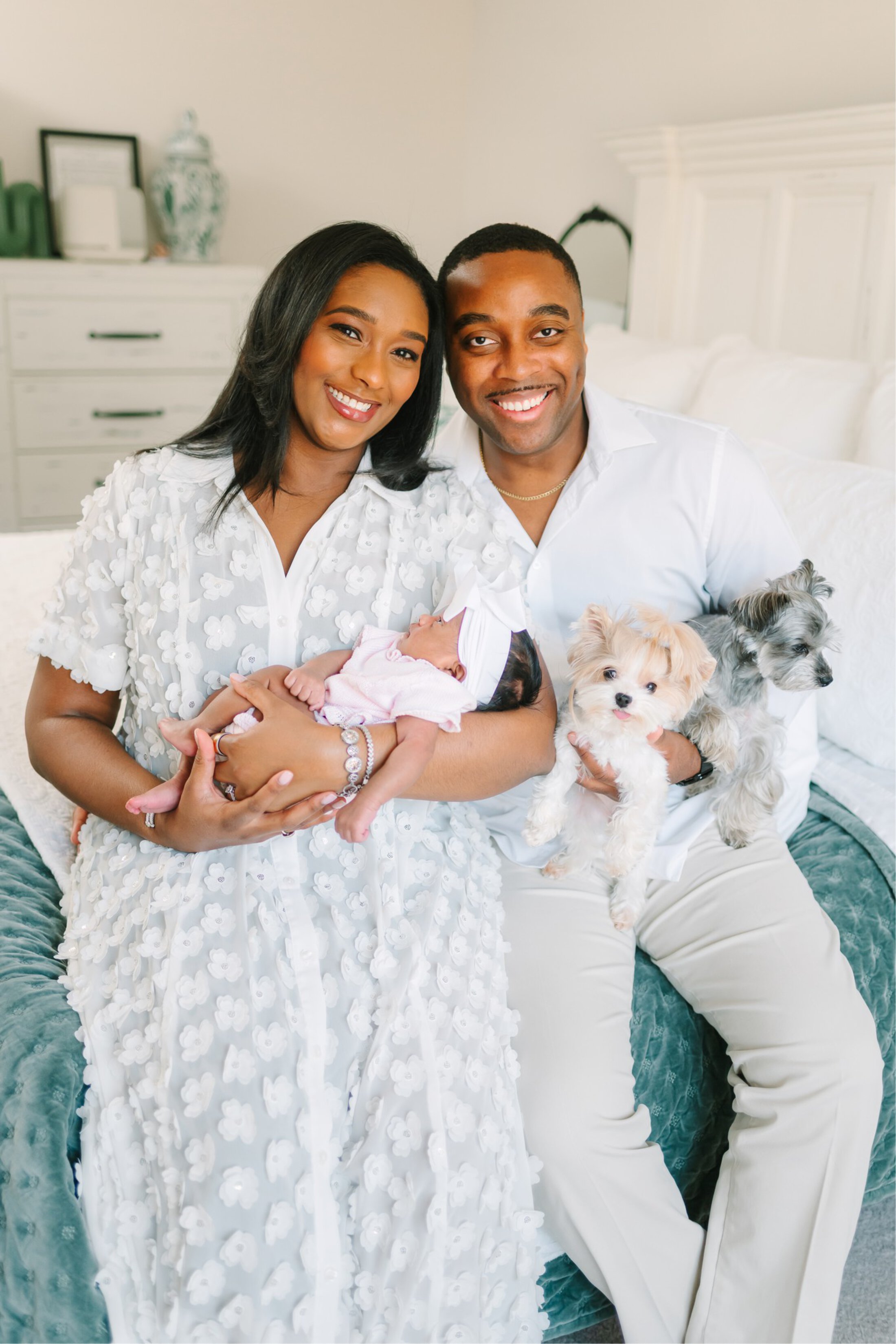 A man and a woman wearing white sit on a bed. She is holding a baby dressed in pale pink. He is holding two small fluffy dogs.