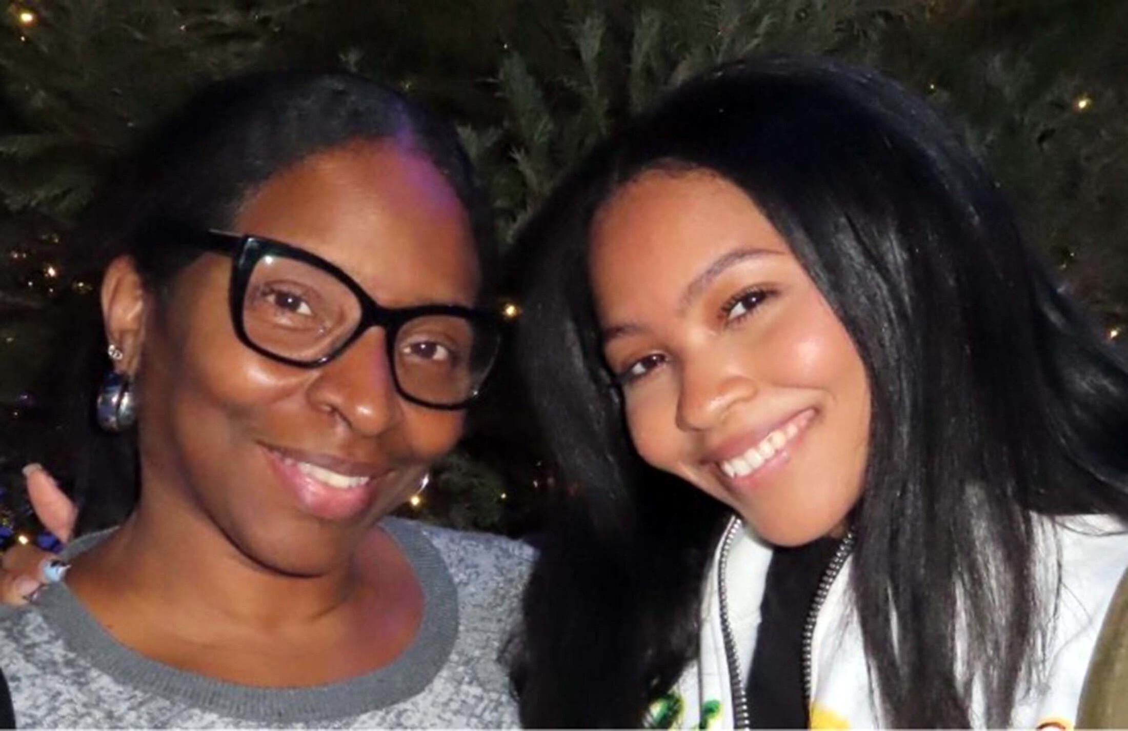 Two women lean toward each other, smiling. The one on the left is wearing glasses and a gray shirt. The one on the right is wearing a white zip-up jacket or vest.