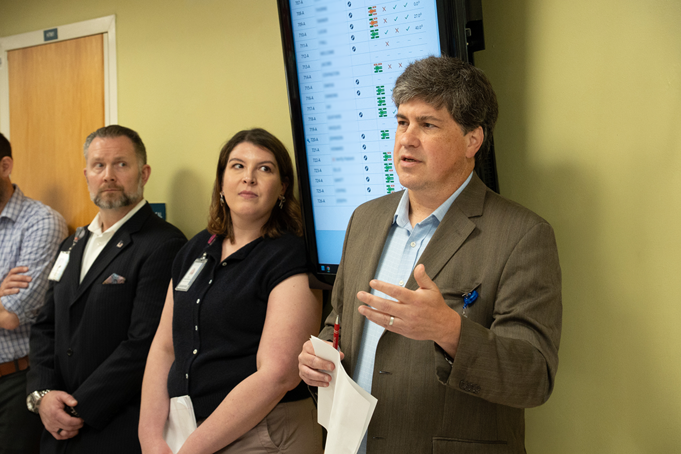 A man addresses a group of professionals in a hospital hallway setting.