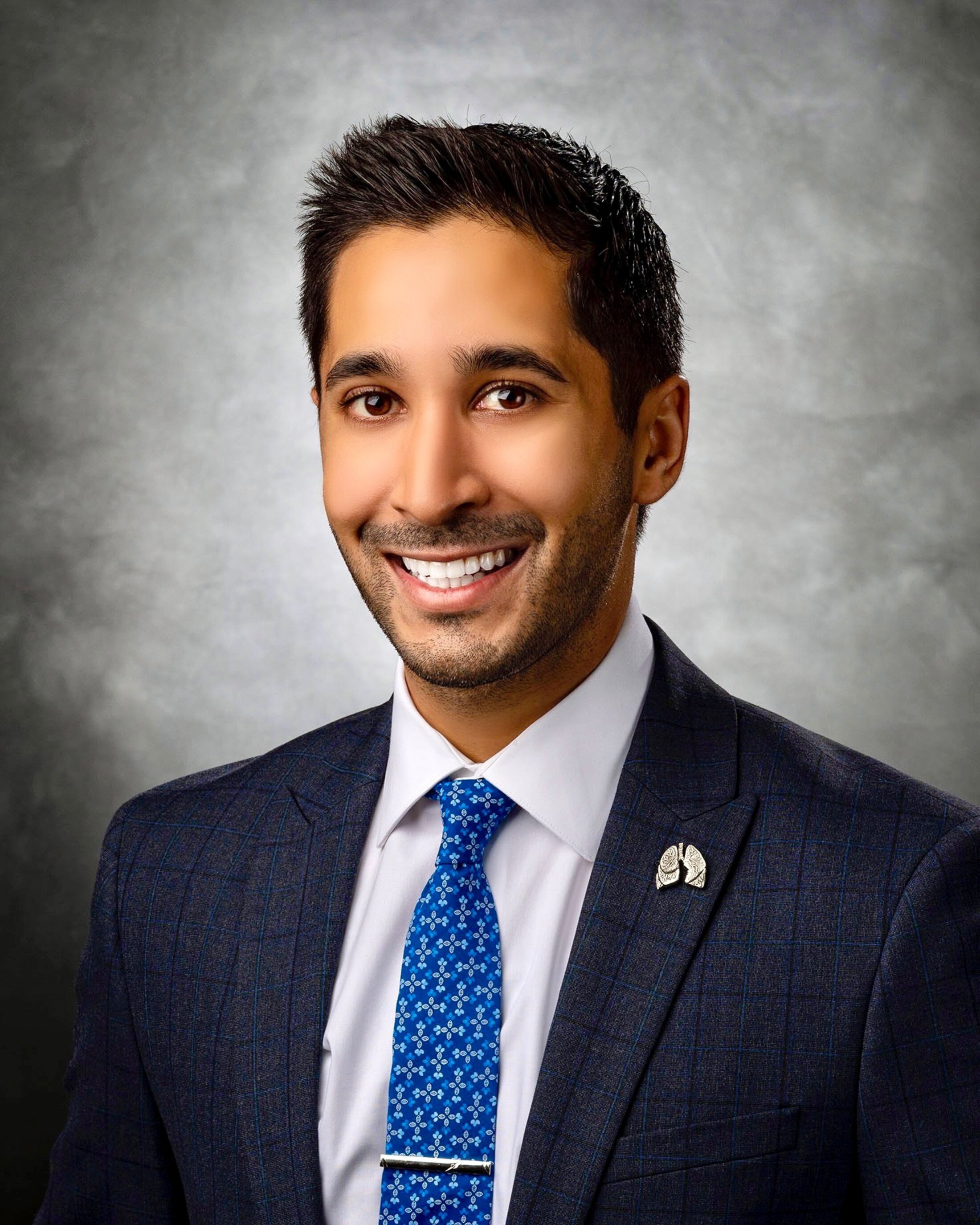 Professional headshot of a smiling man wearing a dark checkered suit, white shirt, and blue patterned tie against a gray studio background
