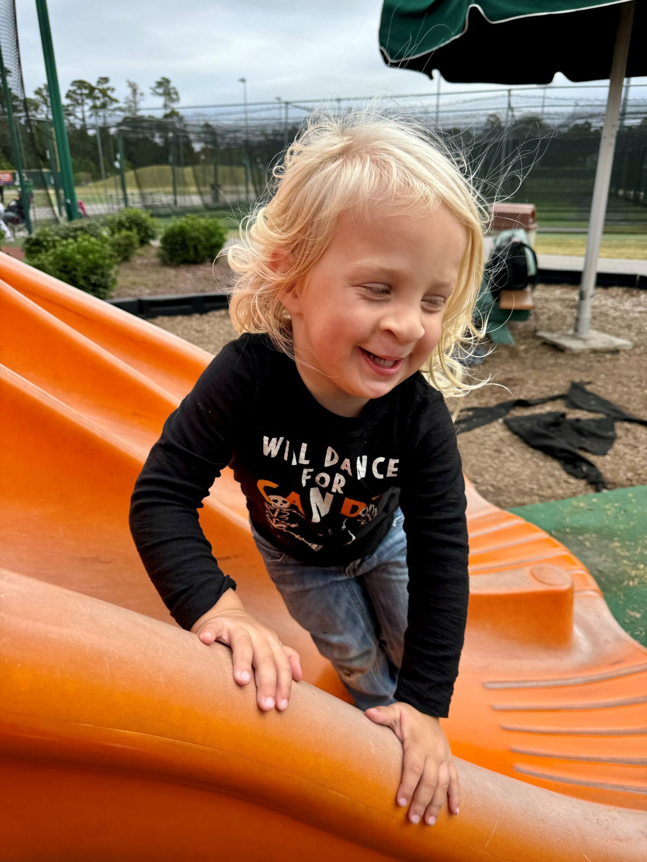 Photograph of a young child climbing an orange playground slide outdoors near tennis courts and greenery. Child wears a black long-sleeve shirt and light blue jeans, with a green canopy and playground equipment visible in the background.